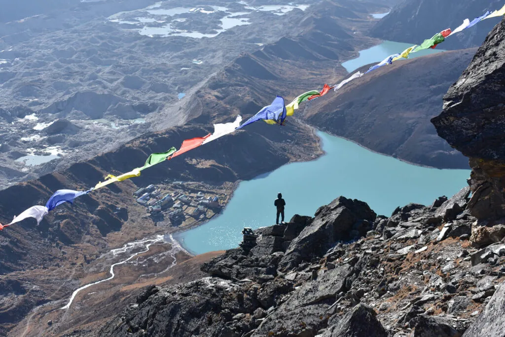 A person stands on a mountain of Everest base camp overlook with a blue lake below. Colourful prayer flags flutter in the wind behind them.